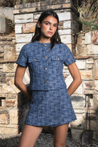 Woman wearing COCO navy blue lurex tweed short sleeve top featuring shimmering textured tweed fabric, subtle metallic lurex detail, round neckline, fitted structured bodice, cropped length, elegant gold point buttons, decorative faux chest pockets, and fully lined construction designed to coordinate with the matching COCO skort.