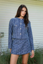Woman wearing COCO navy blue lurex tweed long sleeve jacket featuring textured lurex tweed fabric, structured cropped silhouette, round neckline, clean collarless design, gold tone statement button front closure, decorative chest flap pockets with matching gold tone buttons, subtle metallic threading, and full lining for a smooth finish.