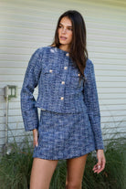 Woman wearing COCO navy blue lurex tweed long sleeve jacket featuring textured lurex tweed fabric, structured cropped silhouette, round neckline, clean collarless design, gold tone statement button front closure, decorative chest flap pockets with matching gold tone buttons, subtle metallic threading, and full lining for a smooth finish.