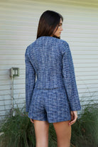 Woman wearing COCO navy blue lurex tweed long sleeve jacket featuring textured lurex tweed fabric, structured cropped silhouette, round neckline, clean collarless design, gold tone statement button front closure, decorative chest flap pockets with matching gold tone buttons, subtle metallic threading, and full lining for a smooth finish.