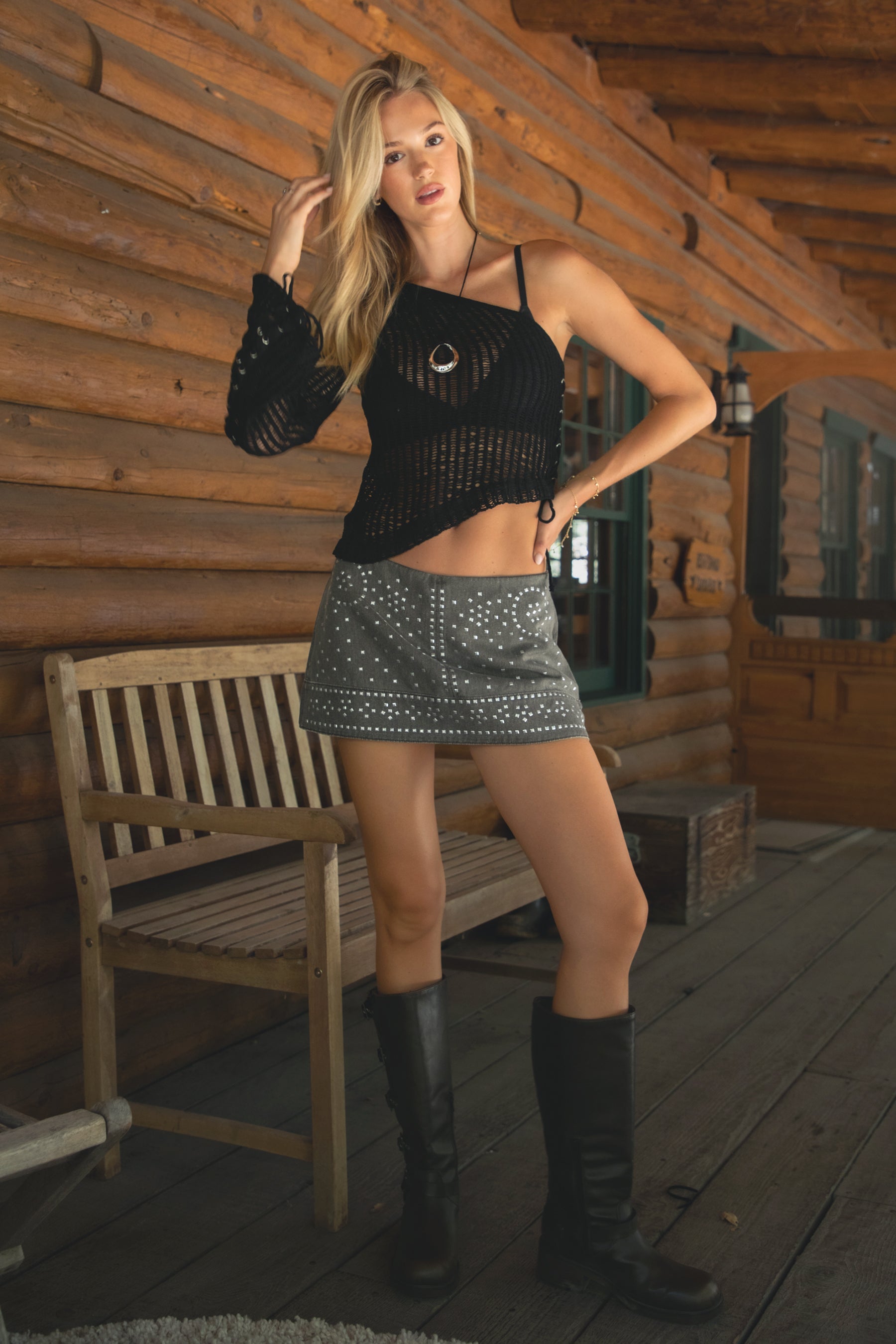Woman wearing TIC TOC DOLCHE Studded Washed Denim Mini Skort in brown featuring built-in undershorts, low rise fit, and allover stud embellishments with ELIEZA One Shoulder Crochet Knit Top in black  and knee high boots on a wooden deck. 