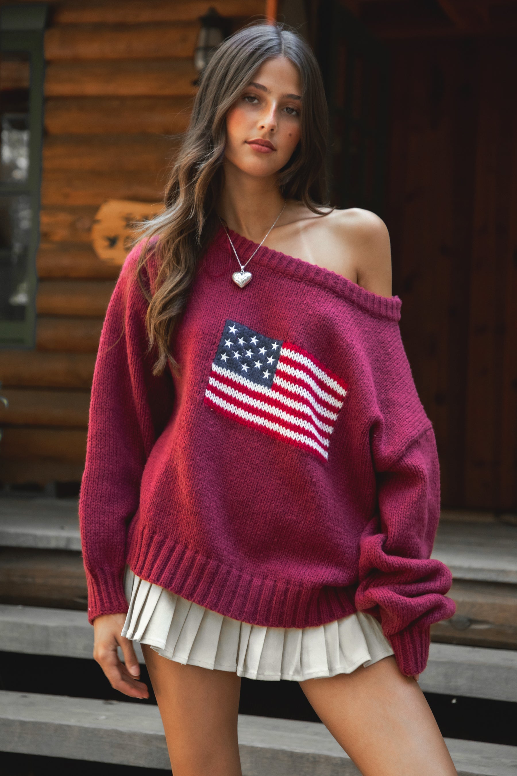 Woman wearing a TIC TOC BRANDI Ribbed Knit Sweater in red with off-the-shoulder boat neck, American flag pattern, and semi-oversized fit, paired with pleated khaki skirt and green rain boots standing on wooden steps.
