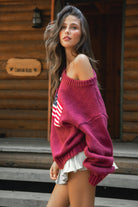 Woman wearing a TIC TOC BRANDI Ribbed Knit Sweater in red with off-the-shoulder boat neck, American flag pattern, and semi-oversized fit, paired with pleated khaki skirt and green rain boots standing on wooden steps.