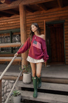 Woman wearing a TIC TOC BRANDI Ribbed Knit Sweater in red with off-the-shoulder boat neck, American flag pattern, and semi-oversized fit, paired with pleated khaki skirt and green rain boots standing on wooden steps.