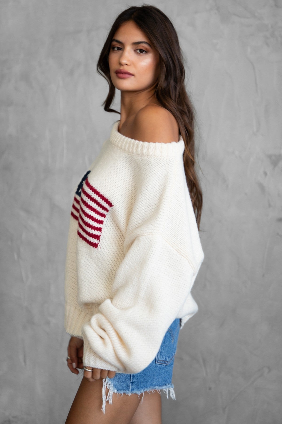 Woman wearing a TIC TOC BRANDI Ribbed Knit Sweater in cream with off-the-shoulder boat neck, American flag pattern, and semi-oversized fit, paired with denim blue shorts.