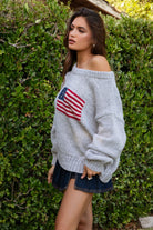 Woman wearing a TIC TOC BRANDI Ribbed Knit Sweater in grey with off-the-shoulder boat neck, American flag pattern, and semi-oversized fit, paired with denim blue skort in front of green bushes.