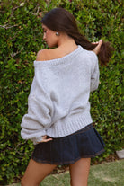 Woman wearing a TIC TOC BRANDI Ribbed Knit Sweater in grey with off-the-shoulder boat neck, American flag pattern, and semi-oversized fit, paired with denim blue skort in front of green bushes.
