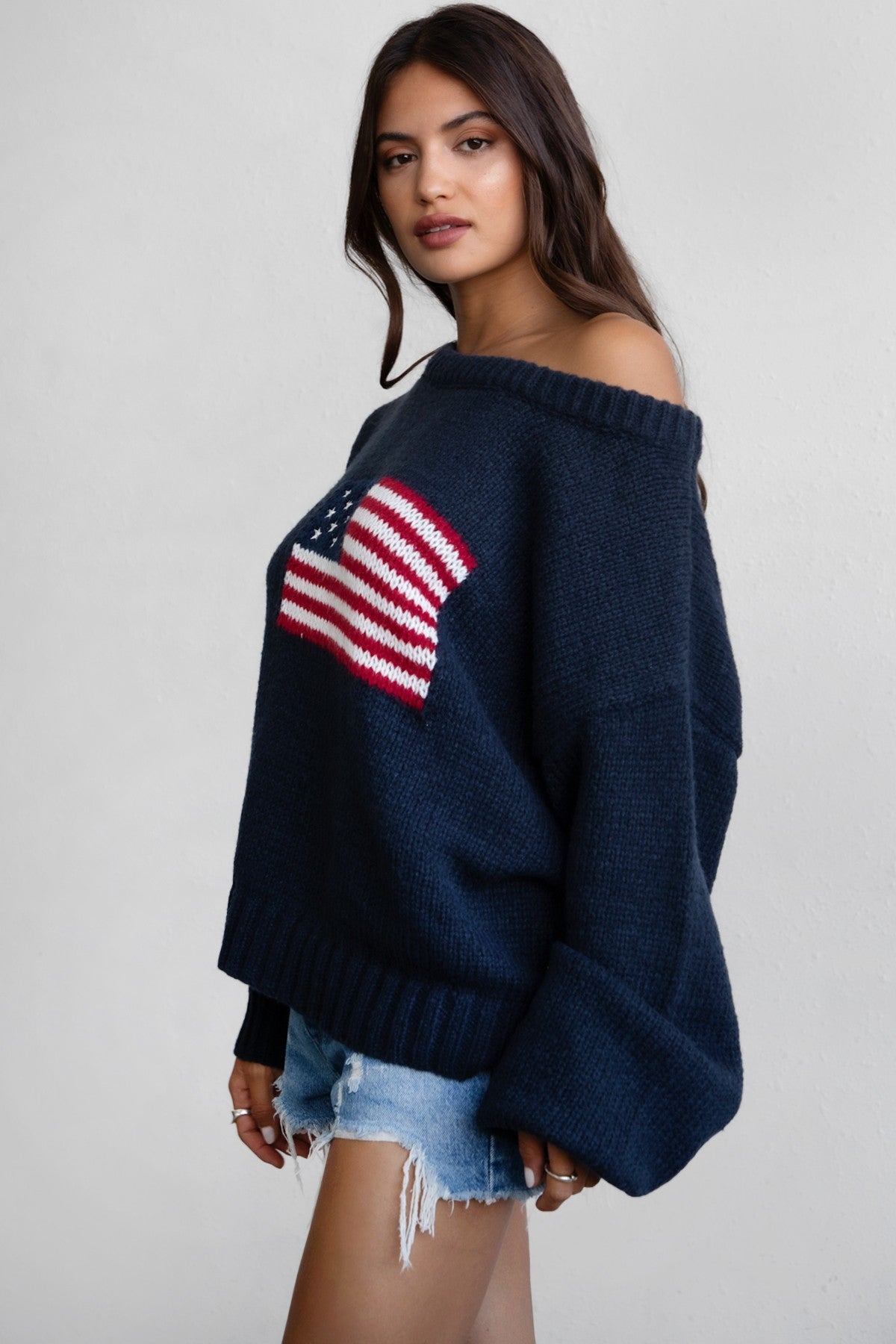 Woman wearing a TIC TOC BRANDI Ribbed Knit Sweater in navy with off-the-shoulder boat neck, American flag pattern, and semi-oversized fit with denim shorts.