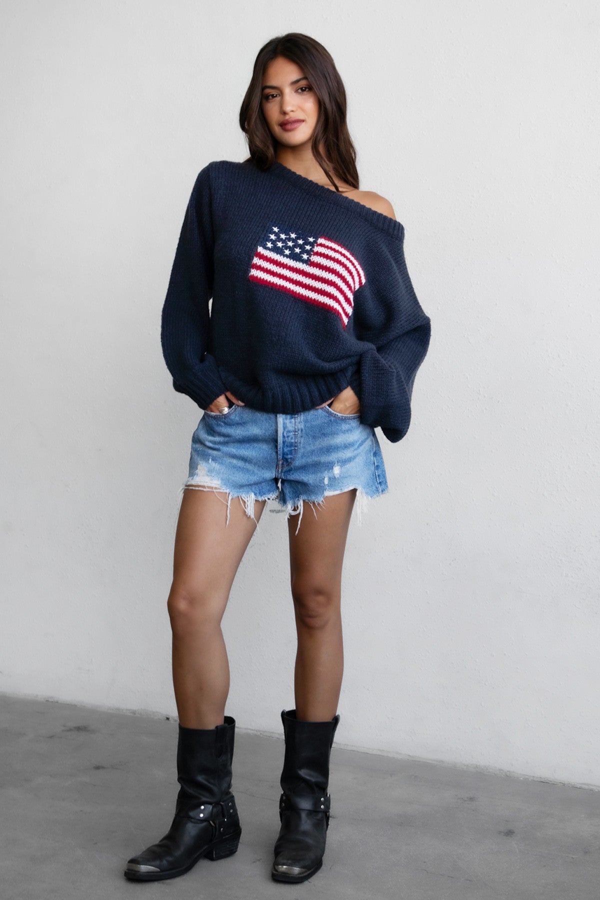 Woman wearing a TIC TOC BRANDI Ribbed Knit Sweater in navy with off-the-shoulder boat neck, American flag pattern, and semi-oversized fit with denim shorts and black boots