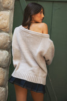 Woman wearing a TIC TOC BRANDI Ribbed Knit Sweater in oatmeal with off-the-shoulder boat neck, American flag pattern, and semi-oversized fit standing against a stone wall and green door.