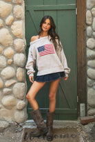 Woman wearing a TIC TOC BRANDI Ribbed Knit Sweater in oatmeal with off-the-shoulder boat neck, American flag pattern, and semi-oversized fit standing against a stone wall and green door.