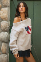 Woman wearing a TIC TOC BRANDI Ribbed Knit Sweater in oatmeal with off-the-shoulder boat neck, American flag pattern, and semi-oversized fit standing against a stone wall and green door.