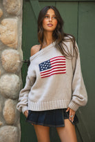 Woman wearing a TIC TOC BRANDI Ribbed Knit Sweater in oatmeal with off-the-shoulder boat neck, American flag pattern, and semi-oversized fit standing against a stone wall and green door.