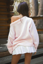 Woman wearing a TIC TOC BRANDI Ribbed Knit Sweater in pink with off-the-shoulder boat neck, American flag pattern, and semi-oversized fit, paired with white denim skirt, standing on wooden steps. 