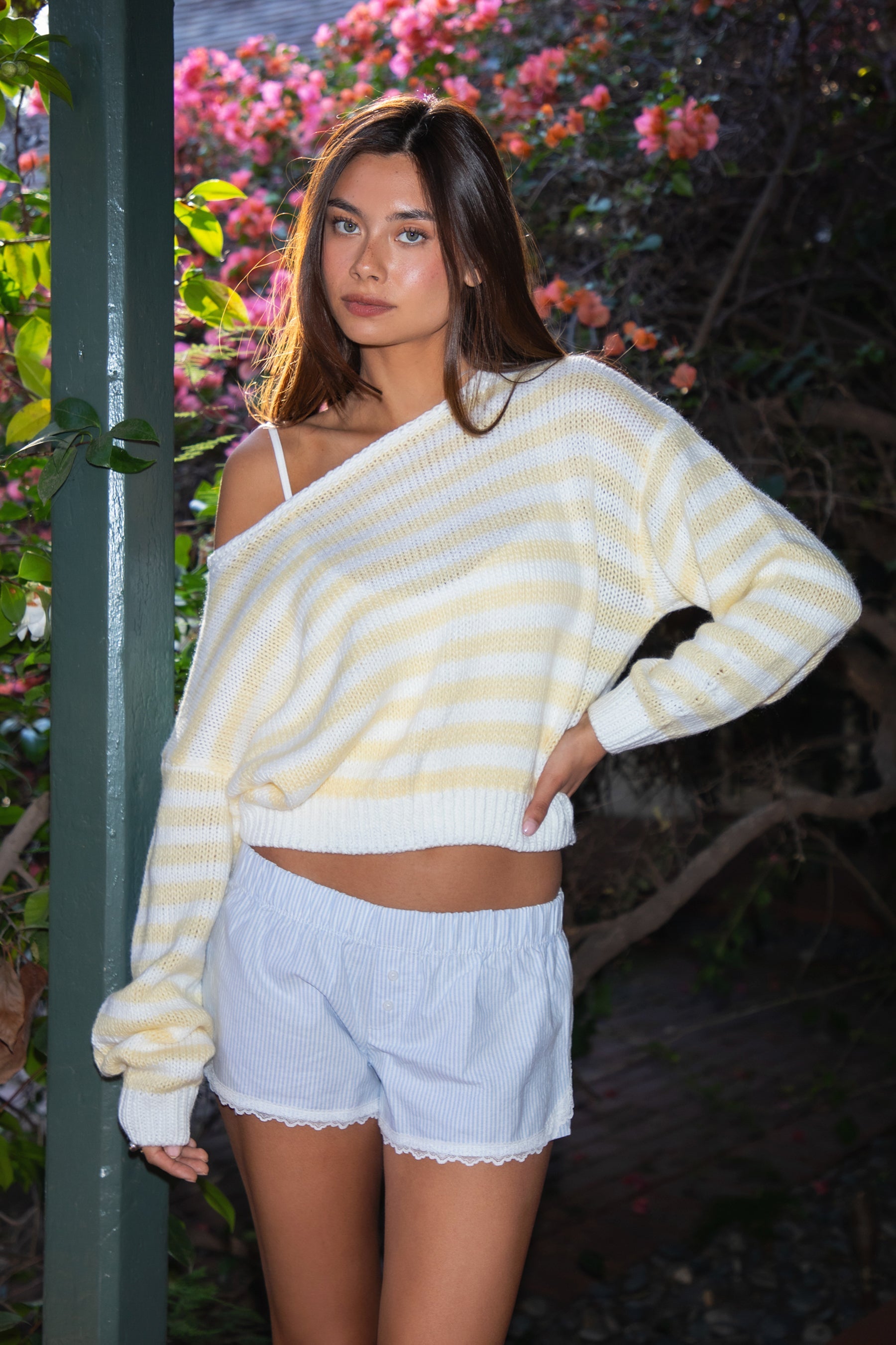 Woman wearing TIC TOC TC31201 PAISEY striped knit off the shoulder oversized sweater with rugby color block pattern inblush and cream paired with a white mini skirt in an outdoor setting. 