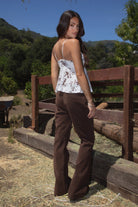 Model wearing the TIC TOC TC31339 ALUA mesh floral babydoll cami top featuring a plunging neckline with lace trim and sheer mesh floral fabric paired with brown suede pants. 