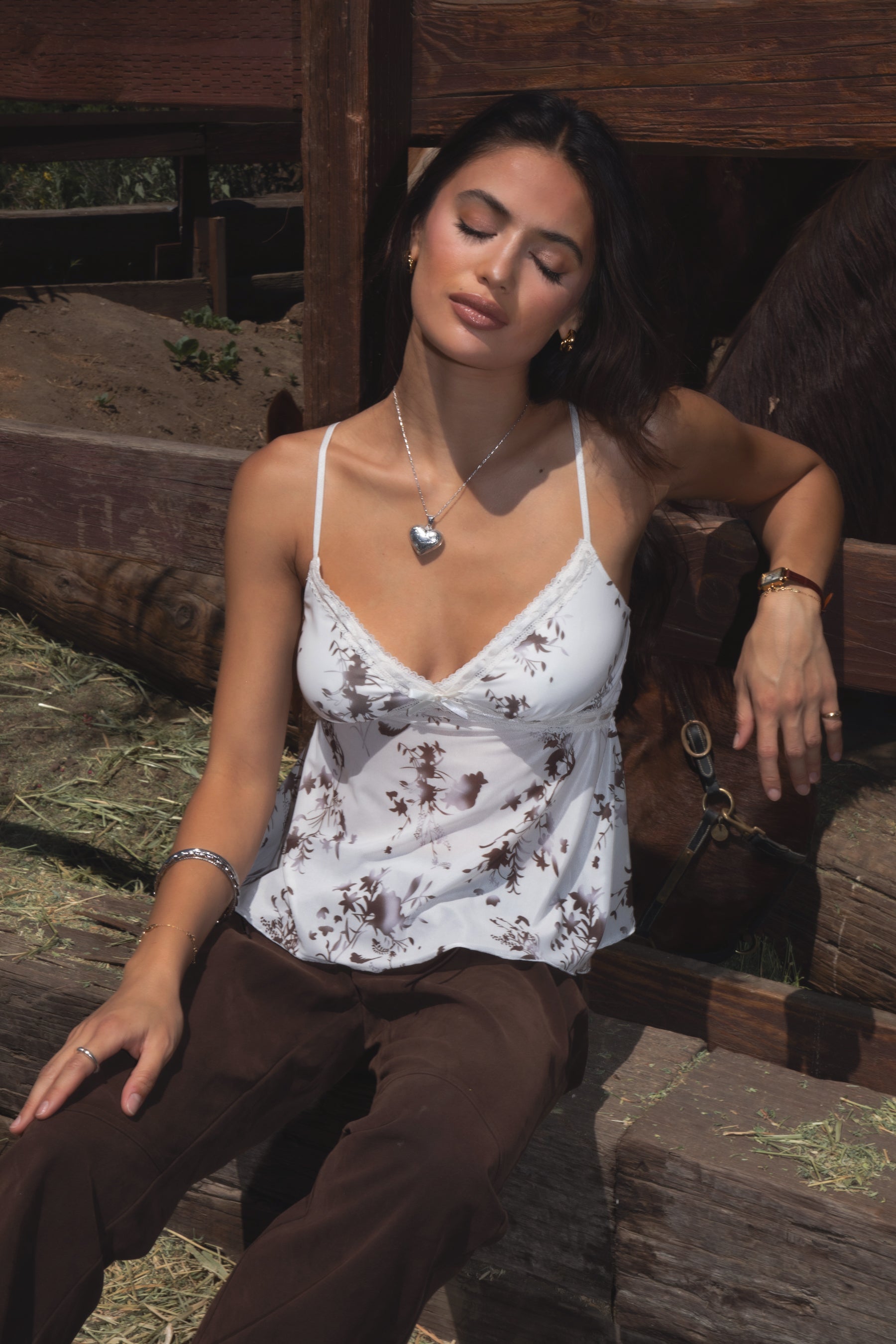 Model wearing the TIC TOC TC31339 ALUA mesh floral babydoll cami top featuring a plunging neckline with lace trim and sheer mesh floral fabric paired with brown suede pants. 