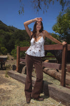 Model wearing the TIC TOC TC31339 ALUA mesh floral babydoll cami top featuring a plunging neckline with lace trim and sheer mesh floral fabric paired with brown suede pants. 