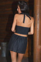 Woman wearing TIC TOC MAEVE Dark Denim Wash Halter Top in dark blue selvedge with plunging neckline, open back, and asymmetrical hem paired with a matching mini skirt against a wooden background
