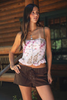 Woman wearing TIC TOC ESME Vintage Inspired Mesh Bustier Corset Top in multi pink featuring lace trim, satin bow, and lace-up front with ruffled details paired with lace up suede shorts in an outdoor cabin setting 