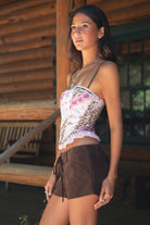 Woman wearing TIC TOC ESME Vintage Inspired Mesh Bustier Corset Top in multi pink featuring lace trim, satin bow, and lace-up front with ruffled details paired with lace up suede shorts in an outdoor cabin setting 