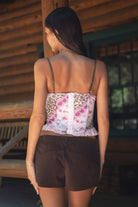 Woman wearing TIC TOC ESME Vintage Inspired Mesh Bustier Corset Top in multi pink featuring lace trim, satin bow, and lace-up front with ruffled details paired with lace up suede shorts in an outdoor cabin setting 