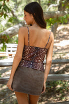 Woman standing outdoors wearing TIC TOC BAMBII Sheer Mesh Deer Print Cami Top in brown with lace trim, adjustable straps, and plunging neckline, paired with a brown denim skirt outdoors with a natural background. 