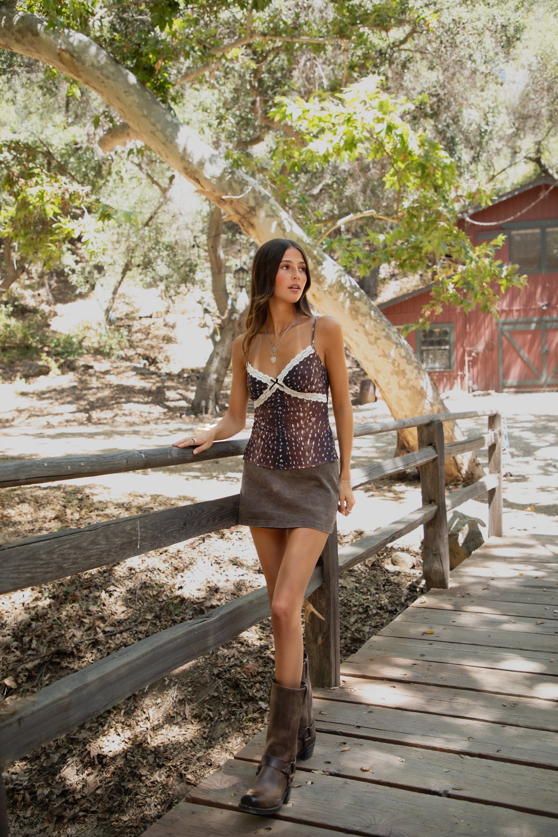 Woman standing outdoors wearing TIC TOC BAMBII Sheer Mesh Deer Print Cami Top in brown with lace trim, adjustable straps, and plunging neckline, paired with a brown denim skirt and brown frye boots, outdoors with a natural background. 