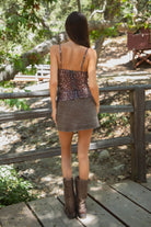 Woman standing outdoors wearing TIC TOC BAMBII Sheer Mesh Deer Print Cami Top in brown with lace trim, adjustable straps, and plunging neckline, paired with a brown denim skirt and brown frye boots, outdoors with a natural background. 