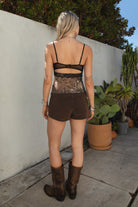 Person wearing a sequin top and brown shorts standing against a white wall with plants in the background.