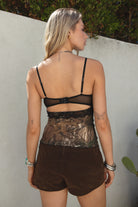 Woman wearing a black lace top and brown shorts against a light-colored wall.