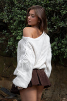 Woman wearing TIC TOC TC31536 BAYLYN ivory off-the-shoulder knit sweater with braided tassel sleeves, cable-knit texture, and relaxed boho fit paired with a brown pleated mini skirt in an outdoor setting.