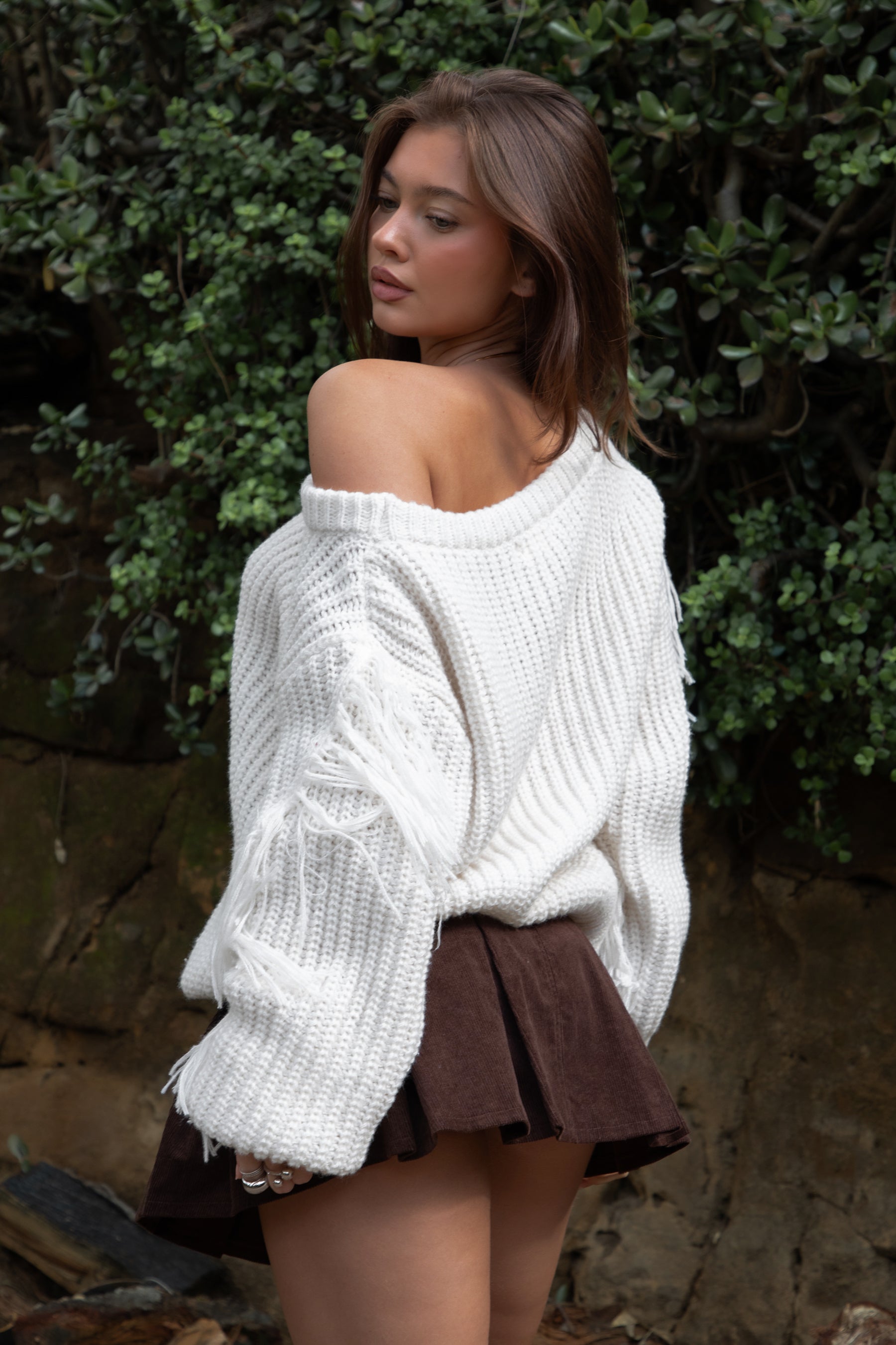 Woman wearing TIC TOC TC31536 BAYLYN ivory off-the-shoulder knit sweater with braided tassel sleeves, cable-knit texture, and relaxed boho fit paired with a brown pleated mini skirt in an outdoor setting.