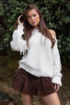 Woman wearing TIC TOC TC31536 BAYLYN ivory off-the-shoulder knit sweater with braided tassel sleeves, cable-knit texture, and relaxed boho fit paired with a brown pleated mini skirt in an outdoor setting.