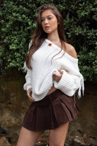 Woman wearing TIC TOC TC31536 BAYLYN ivory off-the-shoulder knit sweater with braided tassel sleeves, cable-knit texture, and relaxed boho fit paired with a brown pleated mini skirt in an outdoor setting.