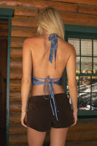 Women wearing TIC TOC INDIRA Mesh Print Halter Cowl Top in blue with self-tie neckline, beaded built-in bra, and soft draped mesh for festival looks paired with brown suede shorts in a cabin setting