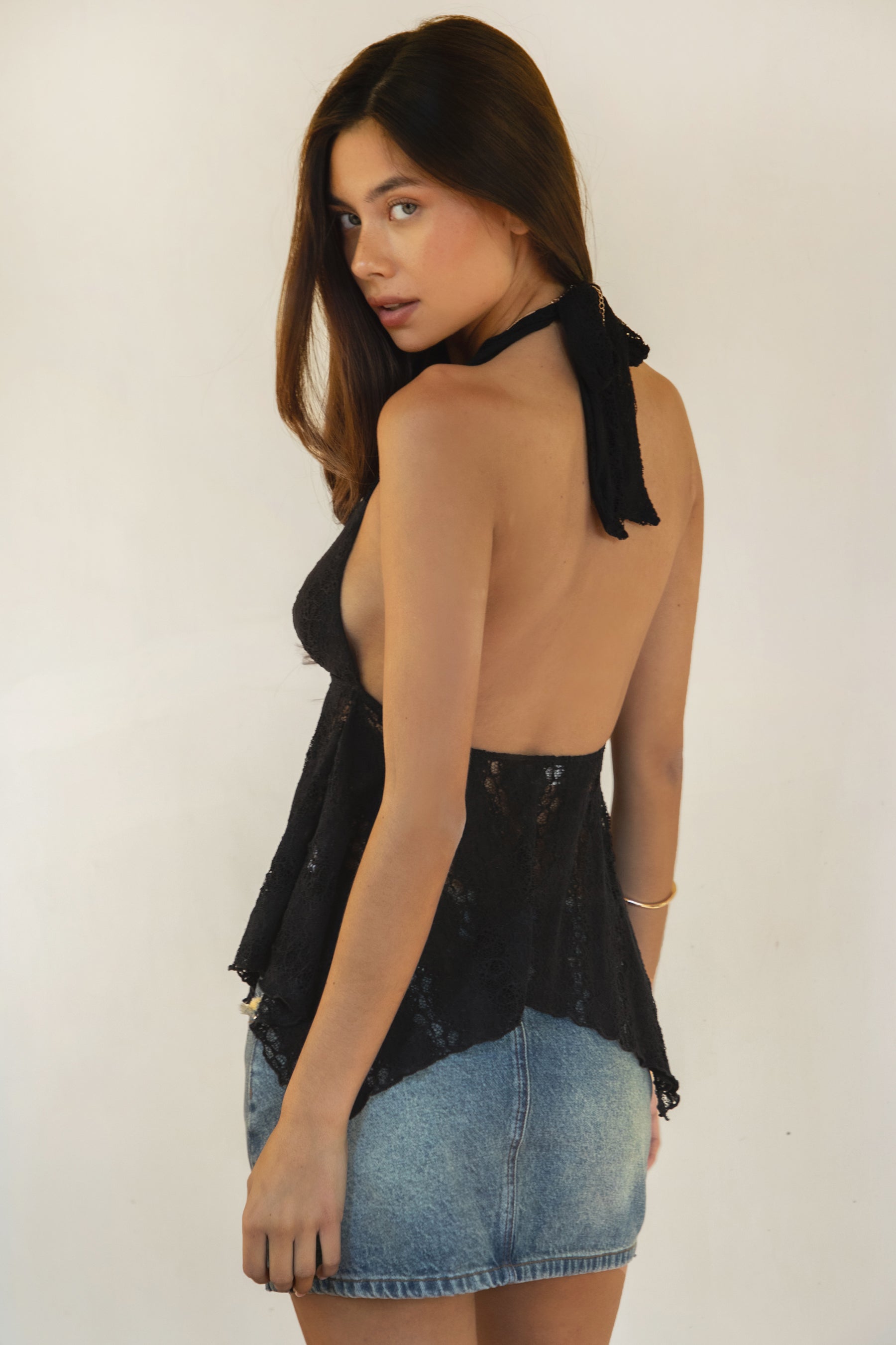 Woman wearing TIC TOC TC31623 LOVELLE deep V-neck lace halter top in black, featuring a plunging neckline, fitted lined bust, sheer lace body, and asymmetrical handkerchief hem, styled with a vintage wash denim mini skirt for a feminine Y2K-inspired look.
