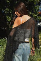Woman wearing TIC TOC TC31650-SOLENA SOLENA crystal sequin crochet poncho featuring lightweight open crochet knit fabric, allover crystal sequin embellishments, a wide off-shoulder neckline, relaxed draped poncho silhouette, cropped asymmetrical hem, and thin tassel fringe detail, styled over a fitted inner layer for a western festival and vacation-ready look.