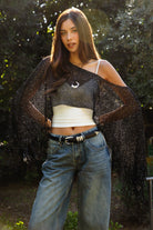 Woman wearing TIC TOC TC31650-SOLENA SOLENA crystal sequin crochet poncho featuring lightweight open crochet knit fabric, allover crystal sequin embellishments, a wide off-shoulder neckline, relaxed draped poncho silhouette, cropped asymmetrical hem, and thin tassel fringe detail, styled over a fitted inner layer for a western festival and vacation-ready look.