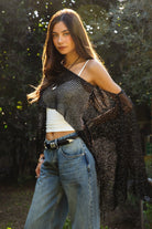 Woman wearing TIC TOC TC31650-SOLENA SOLENA crystal sequin crochet poncho featuring lightweight open crochet knit fabric, allover crystal sequin embellishments, a wide off-shoulder neckline, relaxed draped poncho silhouette, cropped asymmetrical hem, and thin tassel fringe detail, styled over a fitted inner layer for a western festival and vacation-ready look.