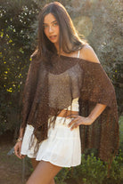 Woman wearing TIC TOC TC31650-SOLENA SOLENA crystal sequin crochet poncho featuring lightweight open crochet knit fabric, allover crystal sequin embellishments, a wide off-shoulder neckline, relaxed draped poncho silhouette, cropped asymmetrical hem, and thin tassel fringe detail, styled over a fitted inner layer for a western festival and vacation-ready look.