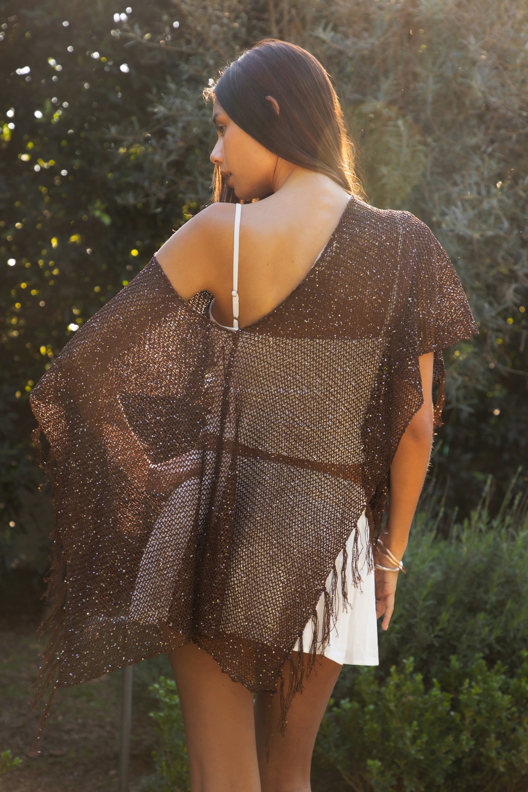 Woman wearing TIC TOC TC31650-SOLENA SOLENA crystal sequin crochet poncho featuring lightweight open crochet knit fabric, allover crystal sequin embellishments, a wide off-shoulder neckline, relaxed draped poncho silhouette, cropped asymmetrical hem, and thin tassel fringe detail, styled over a fitted inner layer for a western festival and vacation-ready look.