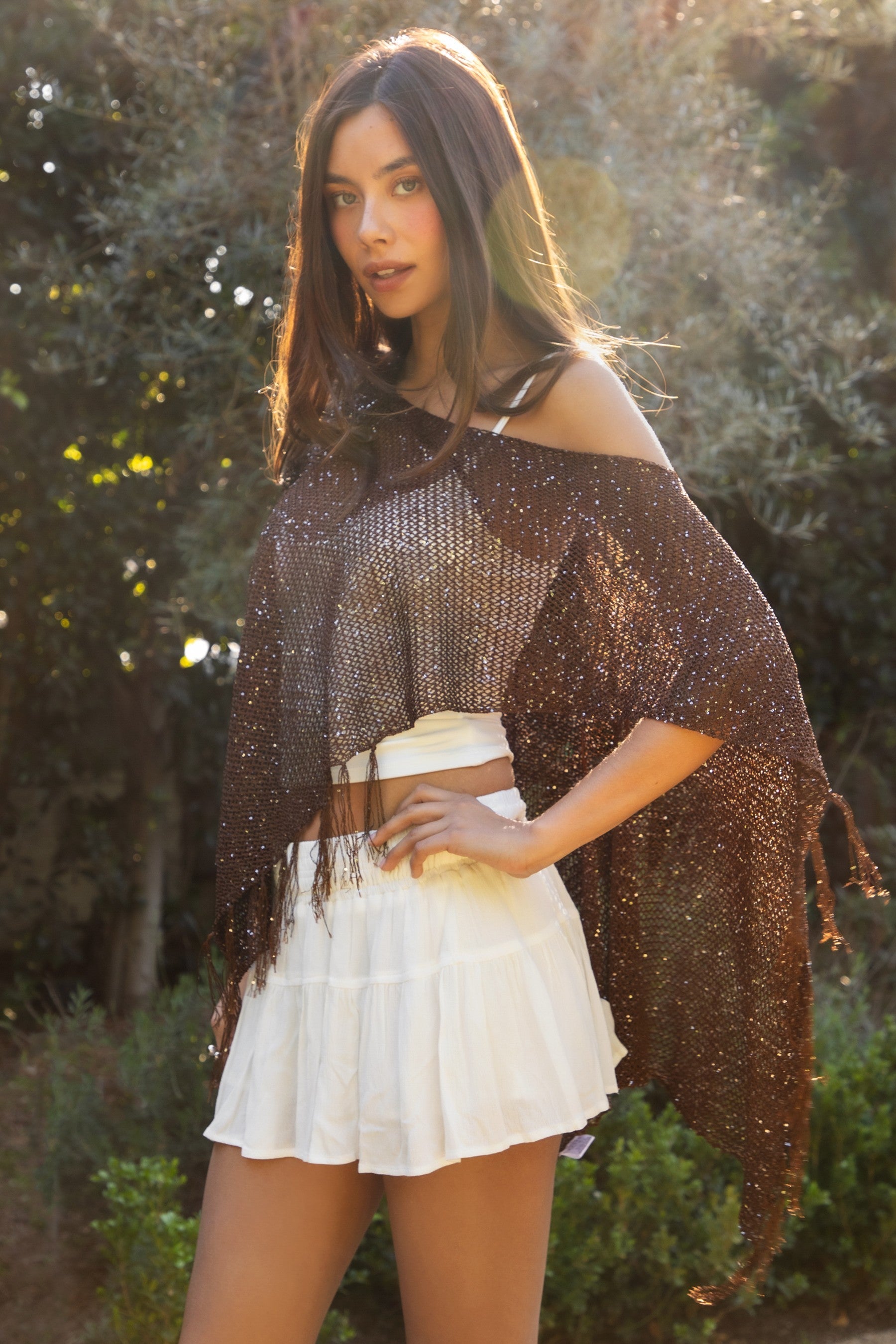 Woman wearing TIC TOC TC31650-SOLENA SOLENA crystal sequin crochet poncho featuring lightweight open crochet knit fabric, allover crystal sequin embellishments, a wide off-shoulder neckline, relaxed draped poncho silhouette, cropped asymmetrical hem, and thin tassel fringe detail, styled over a fitted inner layer for a western festival and vacation-ready look.