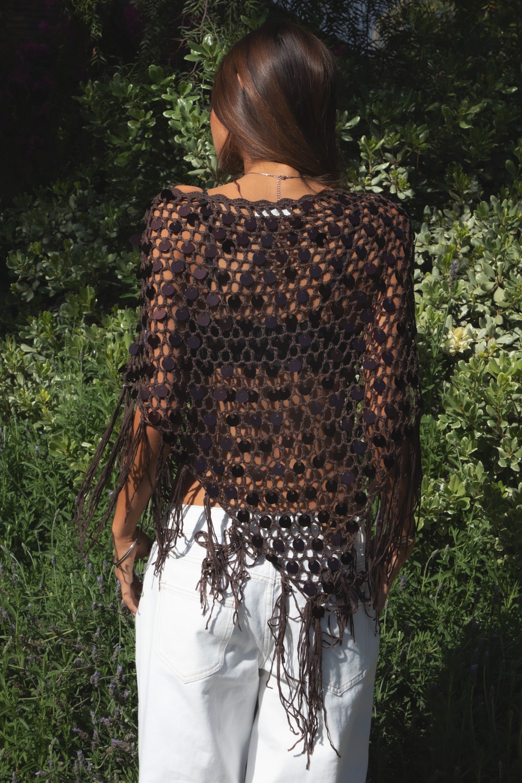 Woman wearing TIC TOC TC31690-LUNARA LUNARA sequin crochet fringe poncho featuring open crochet knit fabric, large allover sequin embellishments, a scalloped wide neckline worn off one shoulder, relaxed poncho silhouette, triangle point hem, and long fringe tassel trim, styled over a fitted top for a western festival and vacation-ready boho look.