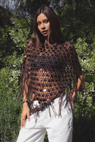 Woman wearing TIC TOC TC31690-LUNARA LUNARA sequin crochet fringe poncho featuring open crochet knit fabric, large allover sequin embellishments, a scalloped wide neckline worn off one shoulder, relaxed poncho silhouette, triangle point hem, and long fringe tassel trim, styled over a fitted top for a western festival and vacation-ready boho look.