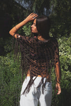 Woman wearing TIC TOC TC31690-LUNARA LUNARA sequin crochet fringe poncho featuring open crochet knit fabric, large allover sequin embellishments, a scalloped wide neckline worn off one shoulder, relaxed poncho silhouette, triangle point hem, and long fringe tassel trim, styled over a fitted top for a western festival and vacation-ready boho look.