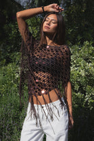 Woman wearing TIC TOC TC31690-LUNARA LUNARA sequin crochet fringe poncho featuring open crochet knit fabric, large allover sequin embellishments, a scalloped wide neckline worn off one shoulder, relaxed poncho silhouette, triangle point hem, and long fringe tassel trim, styled over a fitted top for a western festival and vacation-ready boho look.