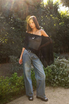 Woman wearing TIC TOC TC31873 SOLEI lightweight open sheer sweater knit poncho in BLACK, featuring a wide off-shoulder neckline, airy open knit texture, relaxed draped poncho silhouette, and asymmetrical hem, styled over a white cami with light wash denim for a western-casual summer layering look.