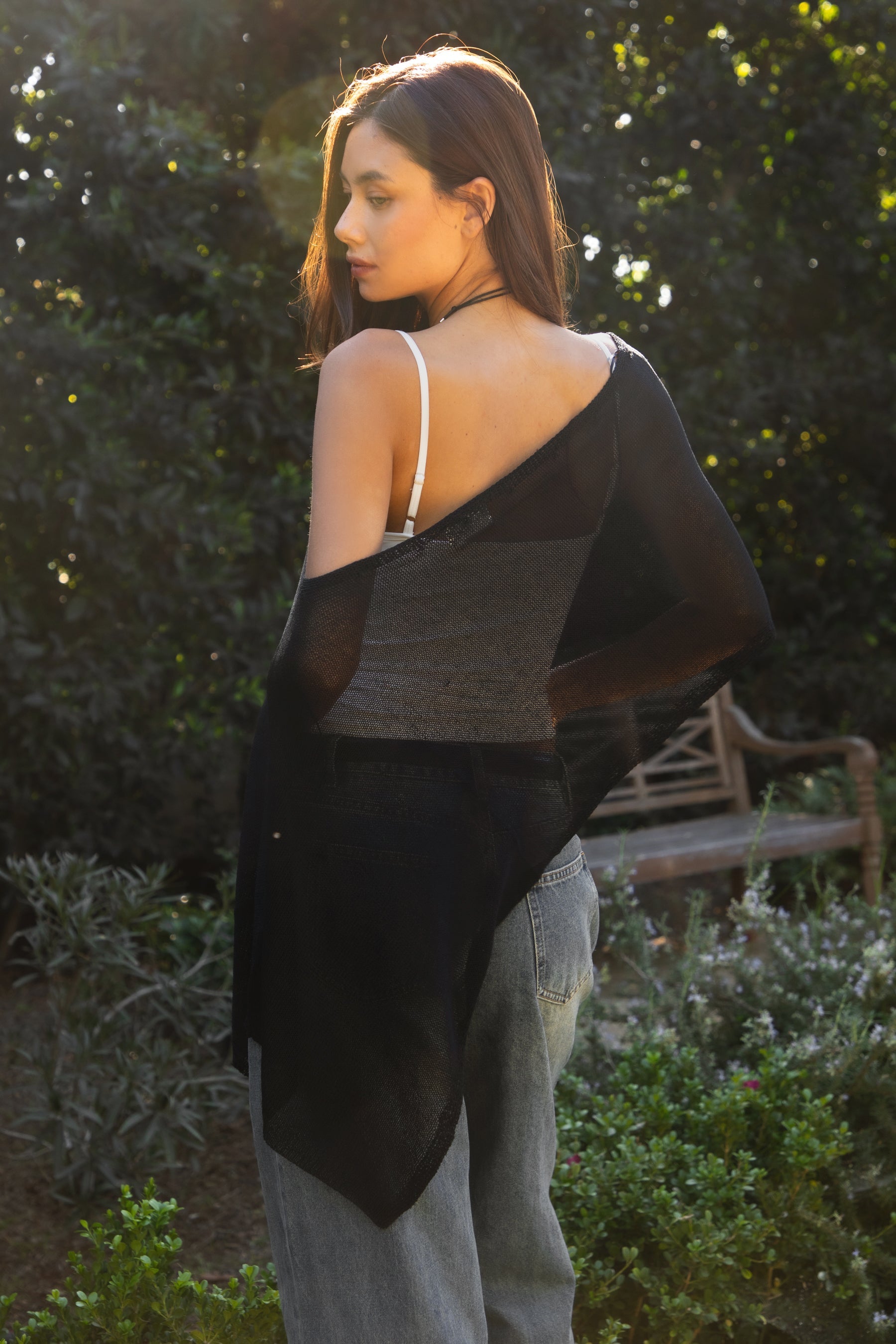 Woman wearing TIC TOC TC31873 SOLEI lightweight open sheer sweater knit poncho in BLACK, featuring a wide off-shoulder neckline, airy open knit texture, relaxed draped poncho silhouette, and asymmetrical hem, styled over a white cami with light wash denim for a western-casual summer layering look.