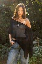 Woman wearing TIC TOC TC31873 SOLEI lightweight open sheer sweater knit poncho in BLACK, featuring a wide off-shoulder neckline, airy open knit texture, relaxed draped poncho silhouette, and asymmetrical hem, styled over a white cami with light wash denim for a western-casual summer layering look.
